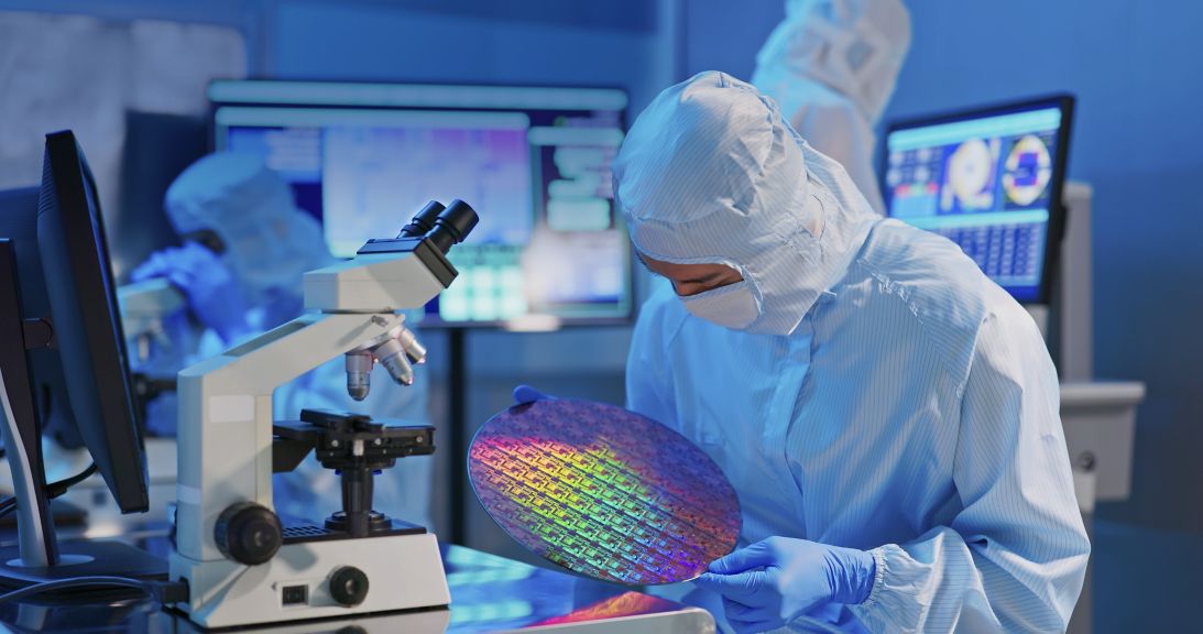 asian male technician in sterile coverall holds wafer that reflects many different colors with gloves and check it at semiconductor manufacturing plant