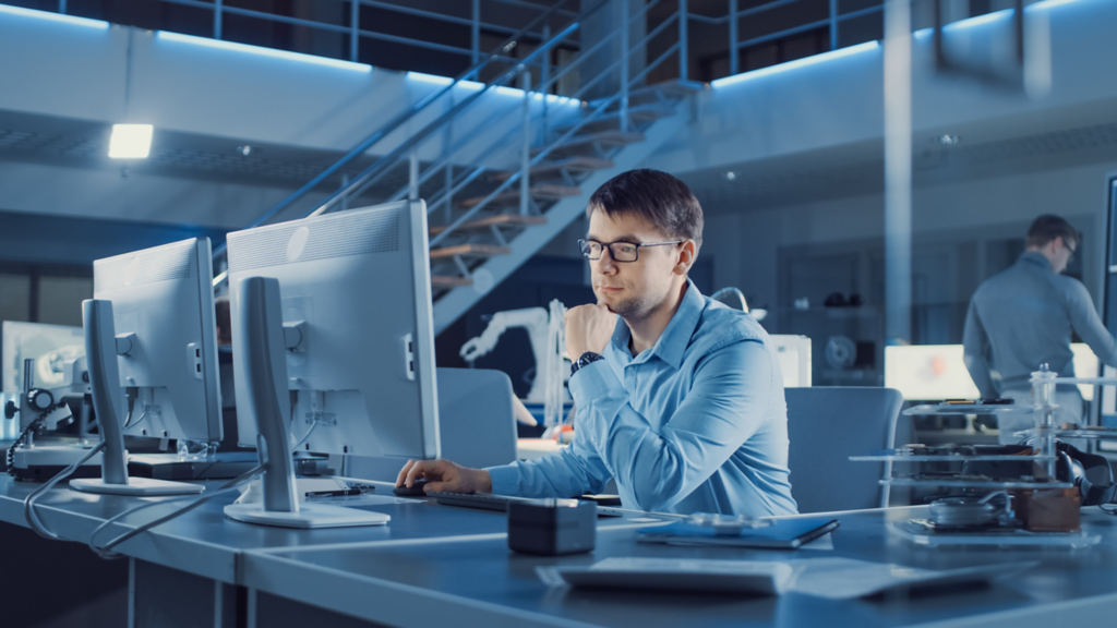 Electronics Development Engineer Sitting at His Desk Uses personal Computer for Programming. Team of Professionals Use Digital Whiteboard with CAD Software for the Modern Industrial Engineering Design